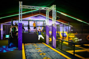  A group of children at an indoor trampoline and adventure park. One child is riding on a zip line over a foam pit while others wait their turn on a platform. The area is brightly lit with neon lighting, and the facility includes various trampoline sections and obstacle course equipment. The slogan "Only if you dare" is displayed on a wall in the background.